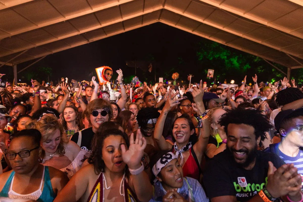 Multidão de pessoas a celebrar numa festa de verão ao ar livre, com bancas e iluminação noturna.