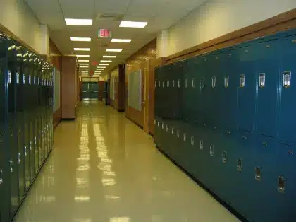 Corredor de escola secundária vazio com folhas de exames sobre uma mesa e figura anónima ao longe