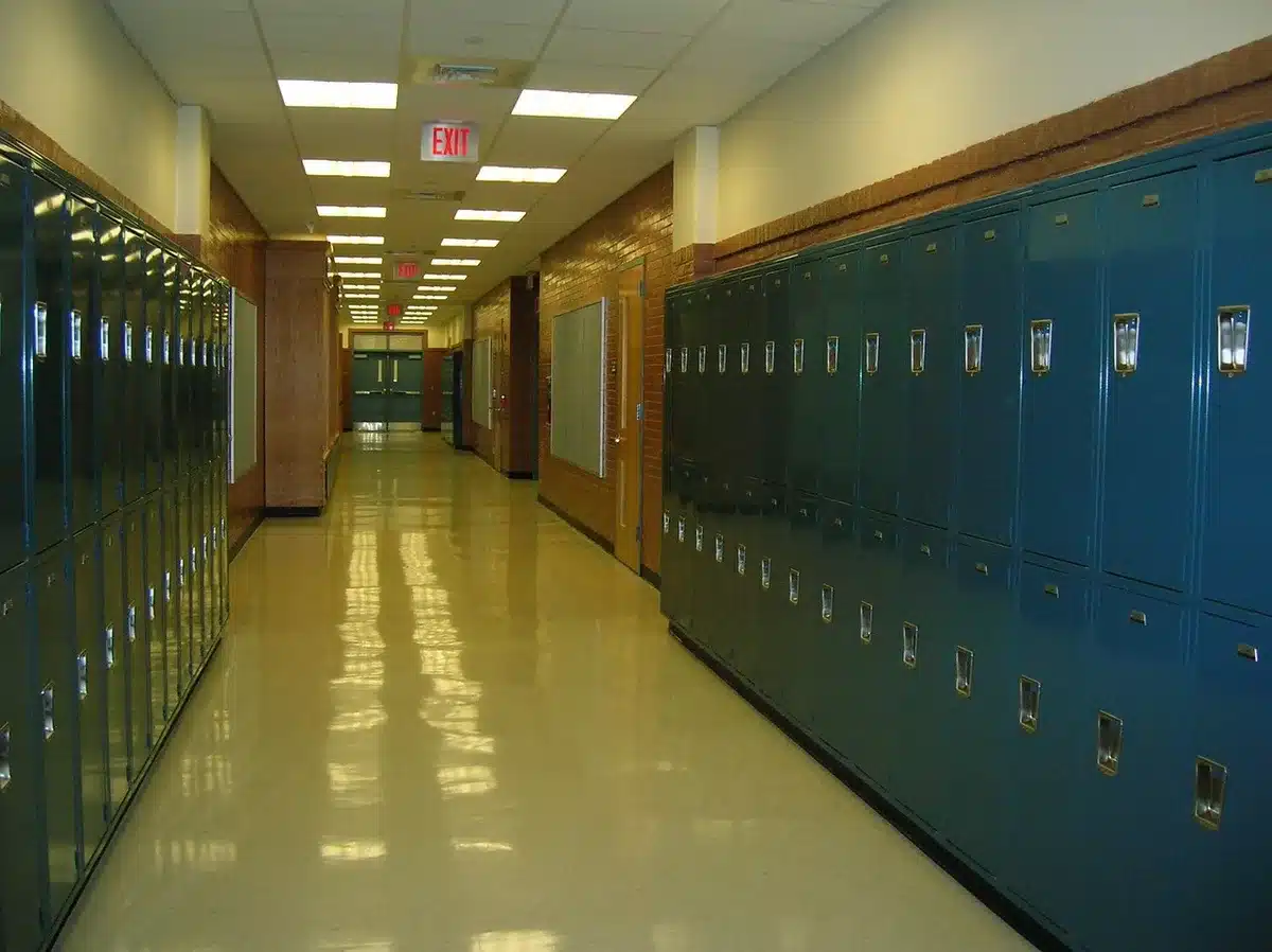 Corredor de escola secundária vazio com folhas de exames sobre uma mesa e figura anónima ao longe