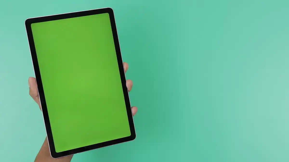 An anonymous person’s hands holding an unbranded tablet at an angle, ambient desk items blurred in background