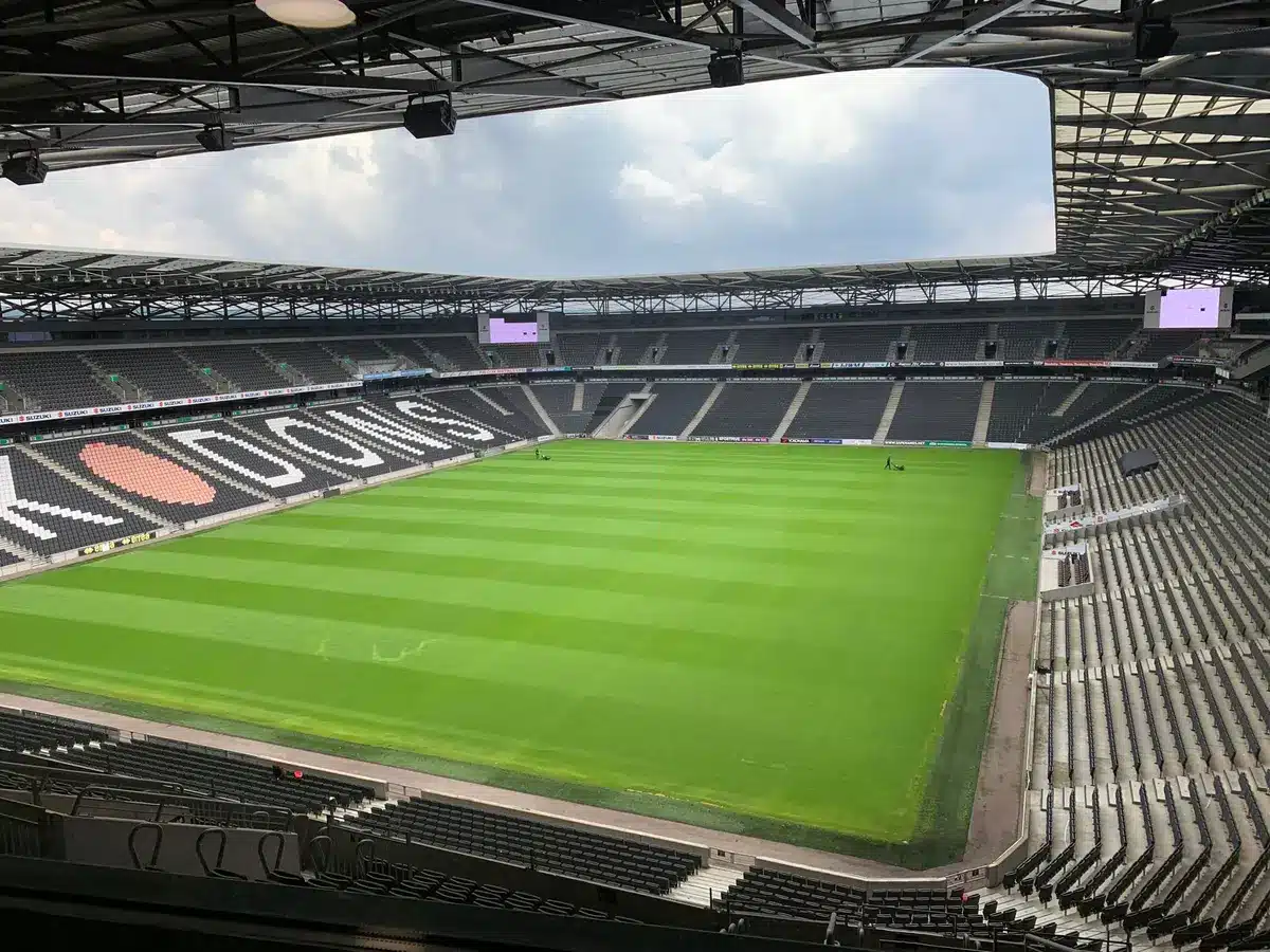 Empty small stadium stand with a single unbranded ball on the pitch, autumn light and distant anonymous figures in the terraces