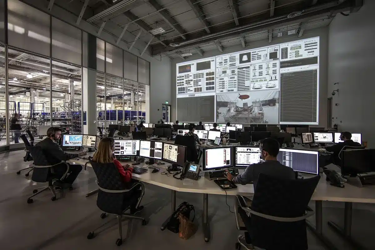 Anonymous silhouette of a person watching multiple screens in a dark control room, screens showing abstract AI-rendered faces and technical readouts, tense atmosphere
