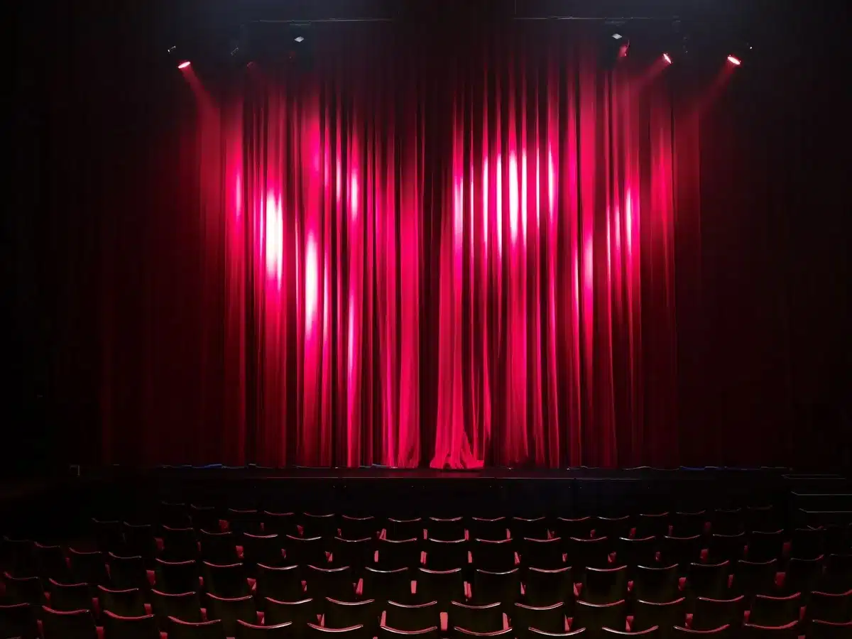 Empty stage with a single microphone under a warm spotlight, an unbranded chair to one side, dust motes visible in the beam